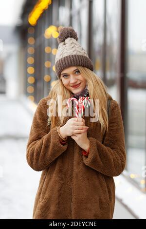 A girl in winter clothes and a hat holds caramel lollipops and candy canes while standing on the street. Shallow focus. Stock Photo