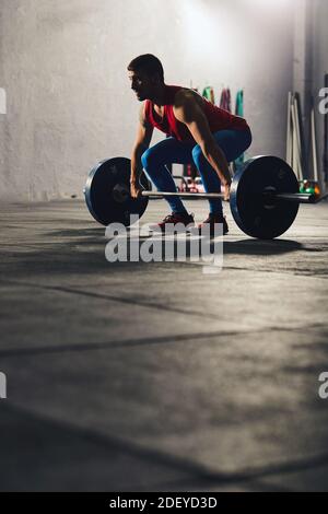 Stock photo of an adult man in a gym squatting down and lifting a barbell. He is wearing sportswear. There are dumbbells in the background. Stock Photo