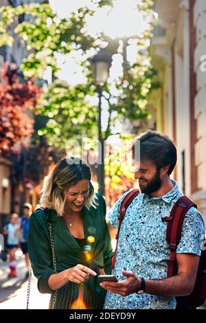 Stock photo of a couple in their 30s walking down a street and looking at a smartphone. They are wearing casual cloth. Stock Photo