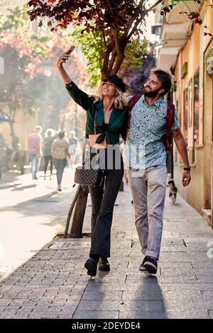 Stock photo of a couple in their 30s walking down a street. They are taking a selfie. They are wearing casual cloth. Stock Photo