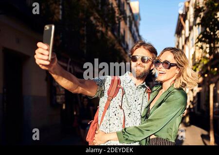 Stock photo of a couple in their 30s walking down a street. They are taking a selfie. They are wearing casual cloth. Stock Photo