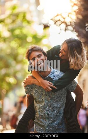 Stock photo of a couple in their 30s. The woman is on the mans back. He is looking at the camera. They are wearing casual cloth. Stock Photo