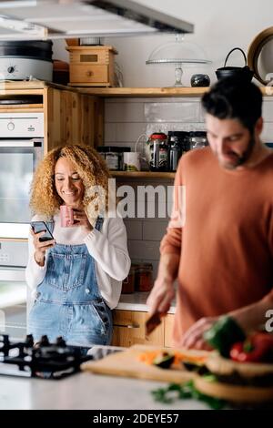 Stock photo of a middle aged woman and man standing in a kitchen. She's using her smartphone and him is cutting vegetables. They are smiling. Stock Photo
