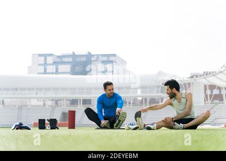 Stock photo of a caucasian and an afro american man sitting down and stretching their legs on a track. They are wearing sportswear. Stock Photo