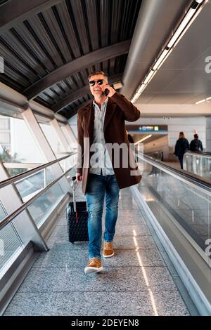 Stock photo of a caucasian business man walking near a belt conveyor with a suitcase going to take a flight. He is smiling and talking through his sma Stock Photo