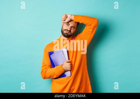 Education. Tired male student holding notebooks and wiping sweat off forehead, looking exhausted, standing in orange sweater against turquoise Stock Photo