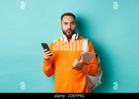 Handsome man student with headphones and backpack, holding digital tablet and smartphone, staring confused at camera, standing against turquoise Stock Photo