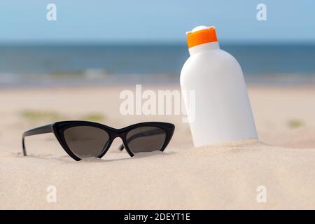 Sunscreen protection lotion and sunglasses in the sand on a sunny day at the beach. White sunblock cream protection bottle from sunburn. Nobody. Stock Photo