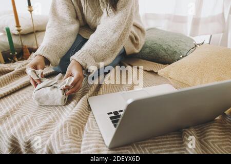 Christmas furoshiki gift wrapping. Female hands wrapping gift in linen fabric close up in festive decorated boho room with laptop, online tutorial. Ze Stock Photo