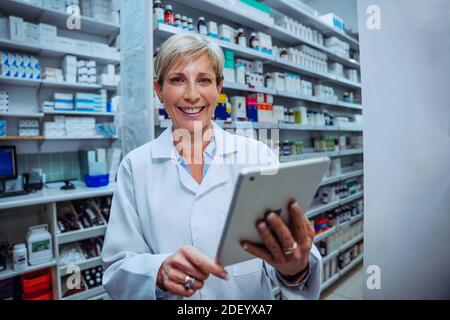 Caucasian senior pharmacist scrolling on digital tablet smiling standing in pharmacy Stock Photo