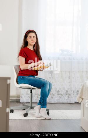 A young Caucasian woman sits beautifully on a chair and poses with a book in her hands. Vertical and copy space. In the background, the white interior Stock Photo