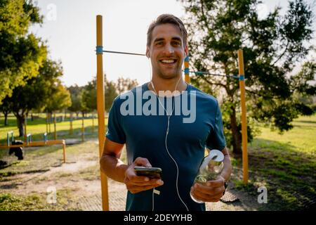 Handsome young male athlete exercising outdoors listening to music in ear phones with smartphone and water bottle Stock Photo