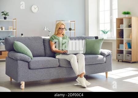 Young woman in eyeglasses sitting on couch and working on project using laptop computer Stock Photo