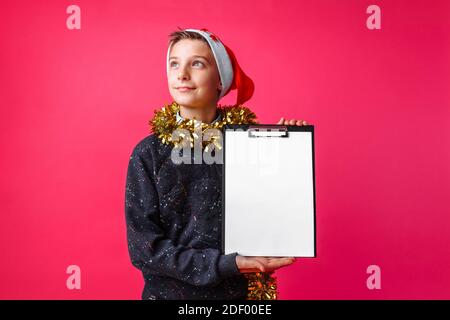 Thoughtful teenager, in Santa hat and with tinsel on his neck, holding a tablet with a clean sheet, the guy makes plans for the new year on a red back Stock Photo