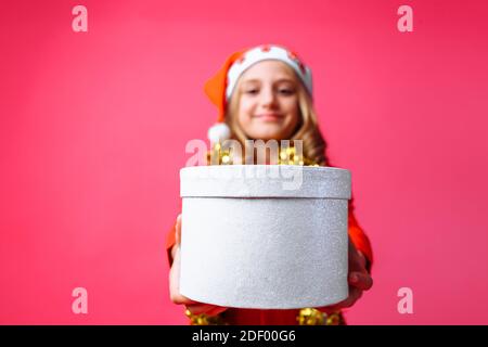 teen girl in Santa's Christmas hat and tinsel around her neck, gives a Christmas gift on a red background. the concept of Christmas to give gifts Stock Photo