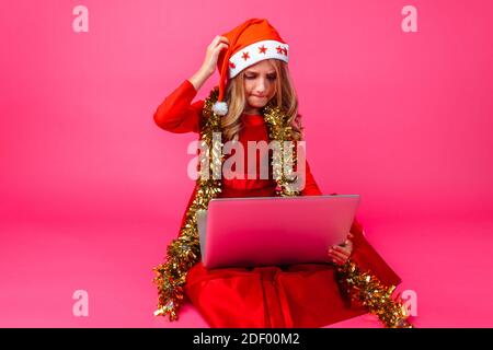 teen girl in Santa hat and tinsel on neck looking at laptop with pensive look posing on red background. The Concept Of Christmas Stock Photo