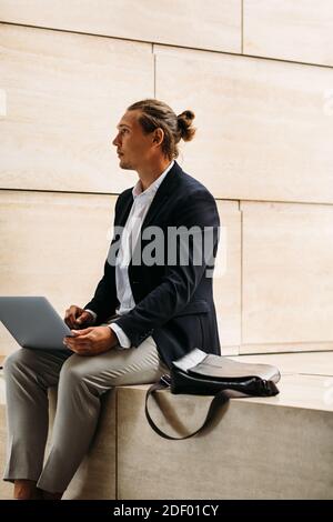 Young businessman sitting outdoors with laptop on his hips looking away Stock Photo
