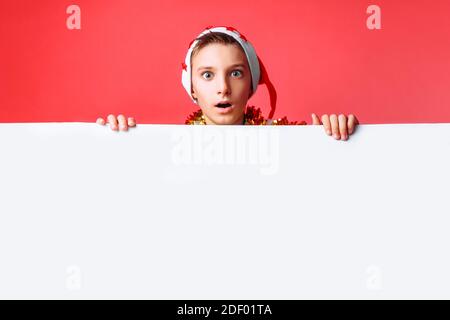 Portrait of a beautiful teen in Santa's hat and with tinsel on her neck, posing behind a white panel isolated on a red background. Teen baby holding a Stock Photo