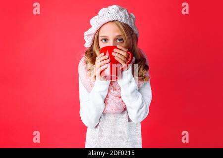 Cute teen girl, basking with a mug of tea, dressed in a pink knitted hat and scarf, on a pink background. Concept, cold, winter, warm clothes Stock Photo