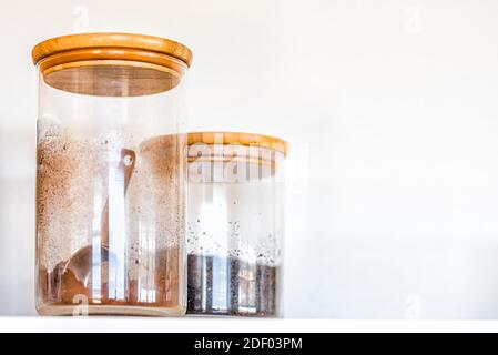 Closeup of two kitchen closed spice glass jars with wooden lids for coffee, cocoa and cinnamon with measuring spoon inside on shelf against white back Stock Photo