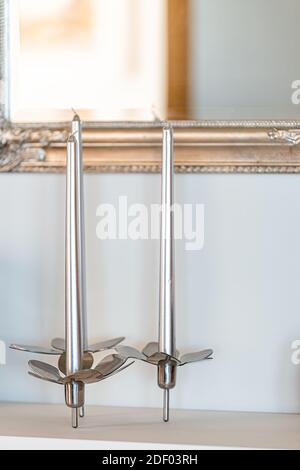 Three modern metal candle sticks stainless steel decorations sculptures of candelabra on table shelf against white wall and mirror in house home Stock Photo