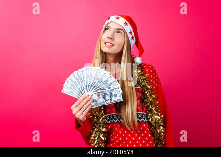 Dreaming girl in a red sweater and a hat of Santa Claus, makes plans and thinks where to spend money, keeps the money on a red background. Christmas, Stock Photo