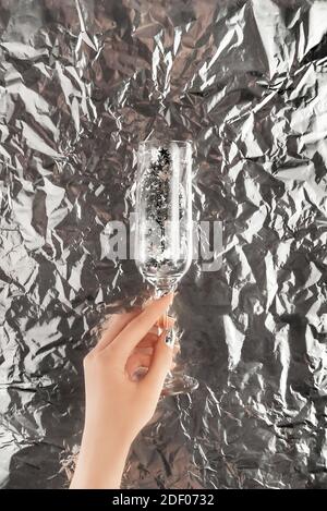 Female hand with trendy silver nail design on foil background holding champagne glass with silver star confetti. Holiday silver manicure. Copy space. Stock Photo
