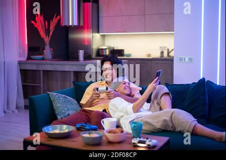 Young woman spending time with her boyfriend at home. She using smart phone while he is playing video games on a gaming console at home. Stock Photo