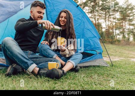 Happiness couple in nature with tent, drinking hot beverage. Man pours tea from thermos to his woman Stock Photo