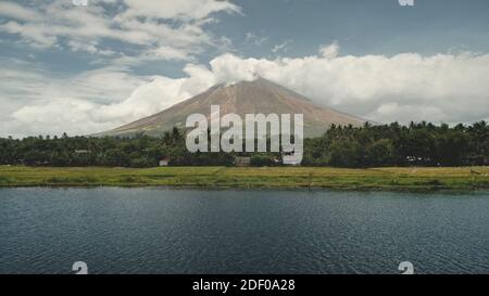 Volcano erupt at green lake shore aerial. Philippines coutryside of Legazpi town at greenery meadows, palm trees. Small cottages at tropic forest. Tropical nobody nature landscape drone shot Stock Photo