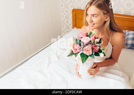 Young beautiful girl with peony flowers, sitting on the bed. She just woke up and was given a bouquet of flowers Stock Photo