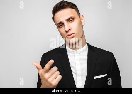 Handsome young business man, male, in a classic black suit, points the finger at the camera, against a gray background. Business concept, career. Stock Photo