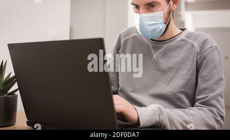 Man remotely working from home using laptop with face mask, looking at screen with virtual web chat. Stock Photo
