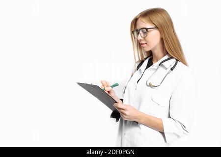 A trainee girl, with glasses, with a stethoscope around her neck, writes the diagnosis in her folder on a white background. medicine concept Stock Photo