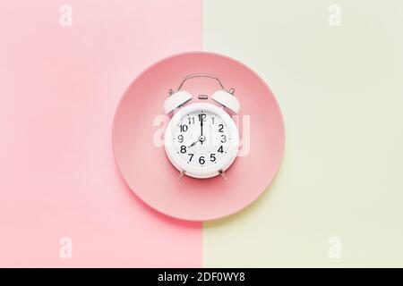 Intermittent fasting concept. White alarm clock on the empty pink plate on the pink-green background. Top view, copy space for text Stock Photo