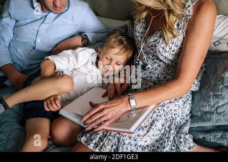 Happy young boy snuggling his parents while they read on bed Stock Photo