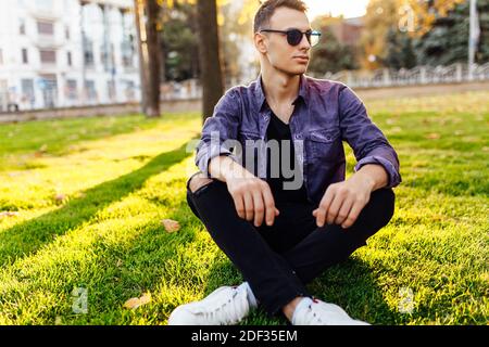 A young man, dressed in casual clothes, sits in a park on the lawn, in sunglasses, at sunset. Stock Photo