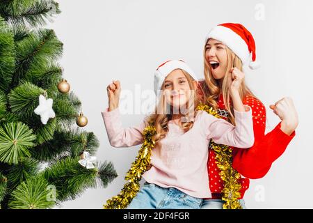 Happy mom and daughter, in Santa's hats, sitting near the Christmas tree, rejoice, showing victory gesture, on white background. Stock Photo