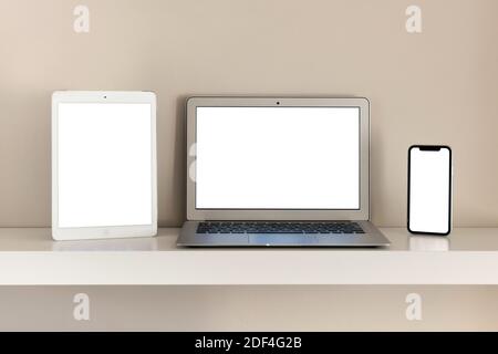 Set of mobile gadgets with blank white screen. Tablet, laptop and smartphone arranged in a row on the shelf against beige wall background, turned on a Stock Photo
