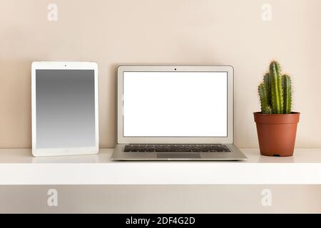 Front view of white tablet and compact laptop notebook with blank screen, sitting on the shelf next to a cactus. Computer monitor copy space concept Stock Photo