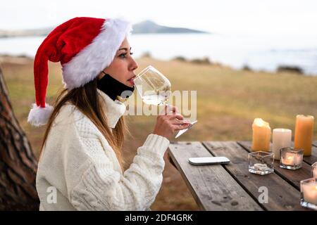 Single beautiful young Caucasian woman drinking white wine wearing Santa Claus hat sitting at a wooden table in the garden of a sea vacation resort wi Stock Photo