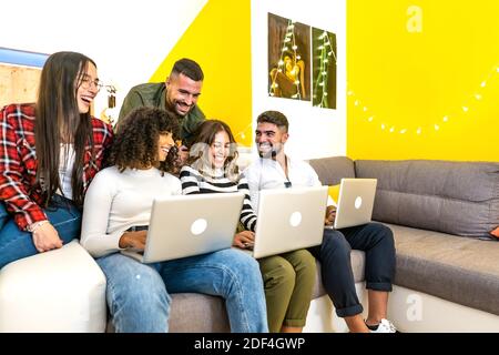 Group of mixed-race friends having fun at home in the living room sitting on the sofa using aluminum laptop - Young people smiling together looking at Stock Photo