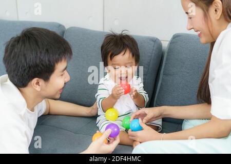 Portrait of Asian cute baby boy playing colorful plastic balls with father and mother on sofa with happy face and smiling to learning colour at home. Stock Photo