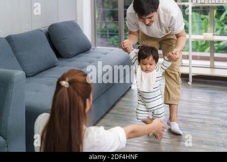 Asian son baby taking first steps walk forward to his mother. Happy little baby learning to walk with father help and teaching how to walk gentlyat ho Stock Photo