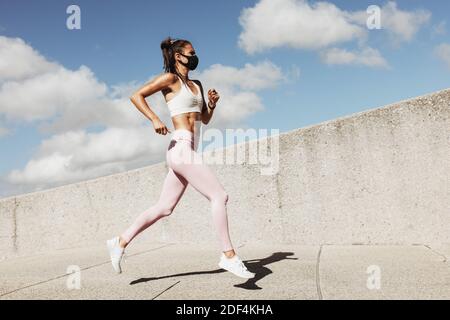 Sports woman wearing face mask running outdoors in the morning. Female athlete in running attire exercising in morning with a face mask on.
