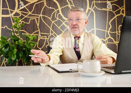 Mature company CEO sitting at office desk with laptop and documents and talking to his employee Stock Photo