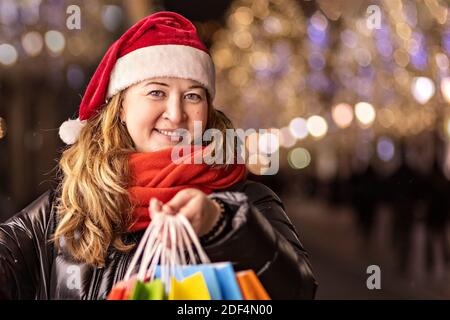 A woman with long hair and a Santa hat near the window of a city store with purchases in colorful, paper bags. New year's shopping. Stock Photo