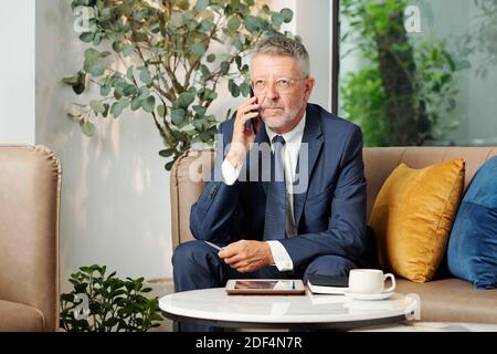 Portrait of serious pensive mature entrepreneur in glasses talking on phone with customer or business partner Stock Photo