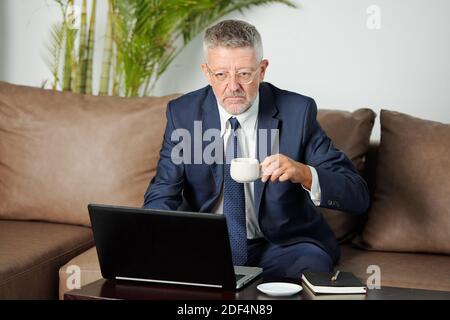 Serious unhappy aged businessman drinking cup of espesso when having morning video phone call with colleagues from company branches Stock Photo