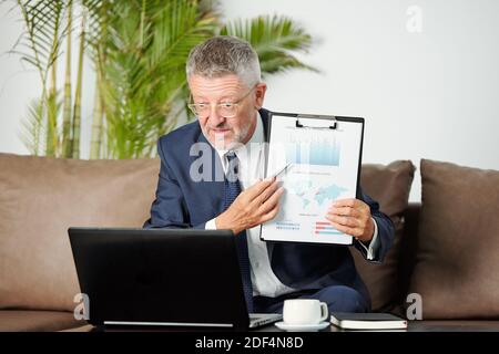 Experinced middle-aged businessman pointing at financial chart when talking to colleague at online confernce on marketing strategy Stock Photo